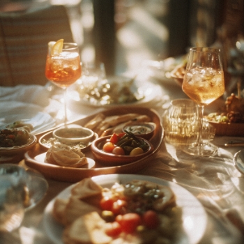 Gedeckter Tisch mit zwei Aperitif-Cocktails in Weingläsern und verschiedenen Schalen mit mediterranen Tapas und Vorspeisen, beleuchtet von warmem Sonnenlicht.