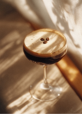 Dunkler Espresso Martini Cocktail in einem Coupette-Glas, garniert mit drei Kaffeebohnen auf der cremigen Schaumkrone, beleuchtet durch Streiflicht und Schattenspiel.