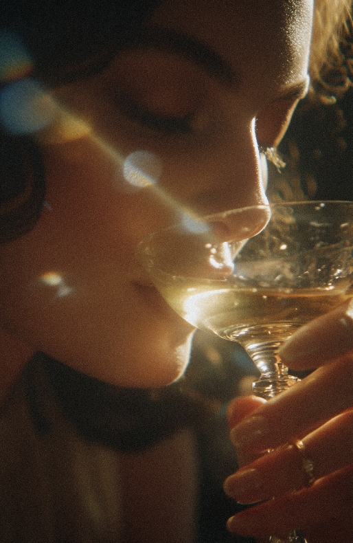 Nahaufnahme einer Frau im Profil, die einen Drink aus einem Vintage-Cocktailglas trinkt. Das Bild hat einen warmen, körnigen Film-Look mit starkem Lichtreflex (Lens Flare) auf dem Glas.