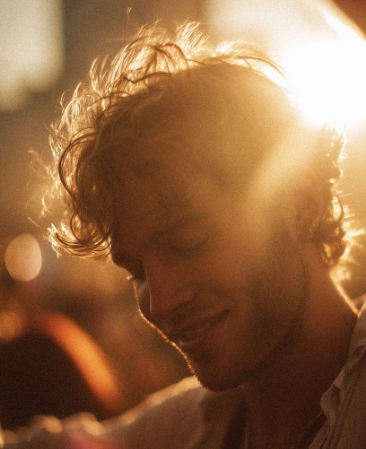 Porträt eines jungen Mannes mit lockigen Haaren, der im warmen Gegenlicht lächelt. Die Aufnahme hat einen körnigen, nostalgischen Film-Look.