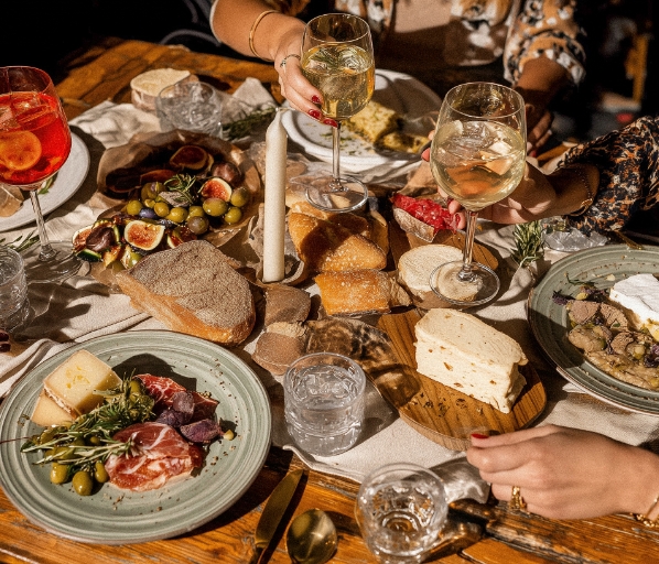 Draufsicht auf einen rustikal gedeckten Tisch mit Käseplatten, Aufschnitt, Brot, Feigen, Oliven, Kerze und Weingläsern mit Weißwein.