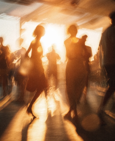 Unscharfe, warme Aufnahme von tanzenden oder sich bewegenden Silhouetten von Menschen in einer hellen, sonnenlichtdurchfluteten Umgebung, mit starkem Gegenlicht-Effekt.