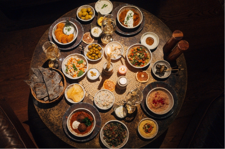 Draufsicht auf einen runden Holztisch, der reich mit einer Auswahl an orientalischen/mediterranen Vorspeisen (Mezze/Tapas) in kleinen Schalen und Gläsern mit Weißwein gedeckt ist, zentriert um eine brennende Kerze.