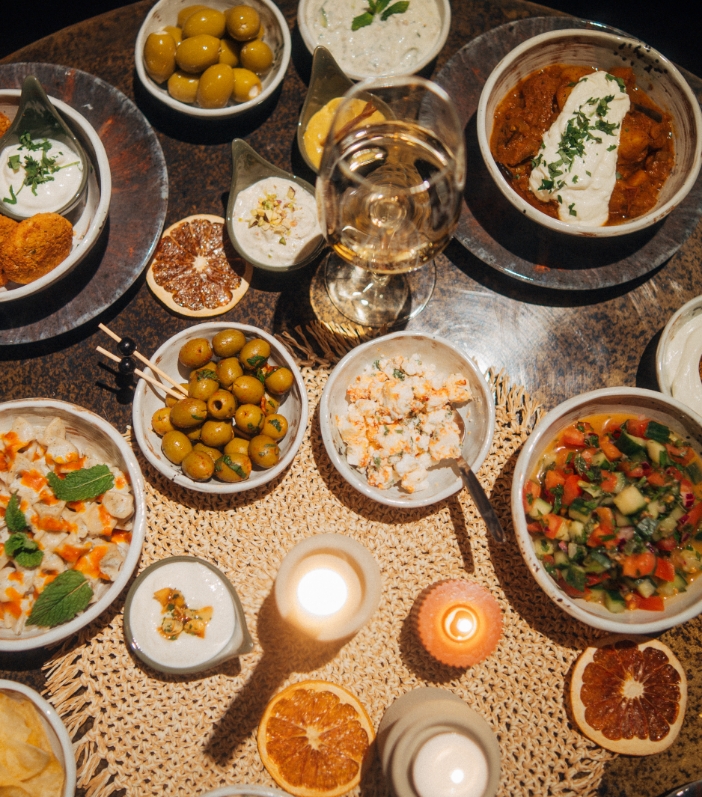 Draufsicht auf einen runden, dunklen Tisch, der reich mit einer großen Auswahl an orientalischen/mediterranen Vorspeisen (Mezze/Tapas) in kleinen Schalen und Gläsern mit Weißwein gedeckt ist, zentriert um eine Kerze.
