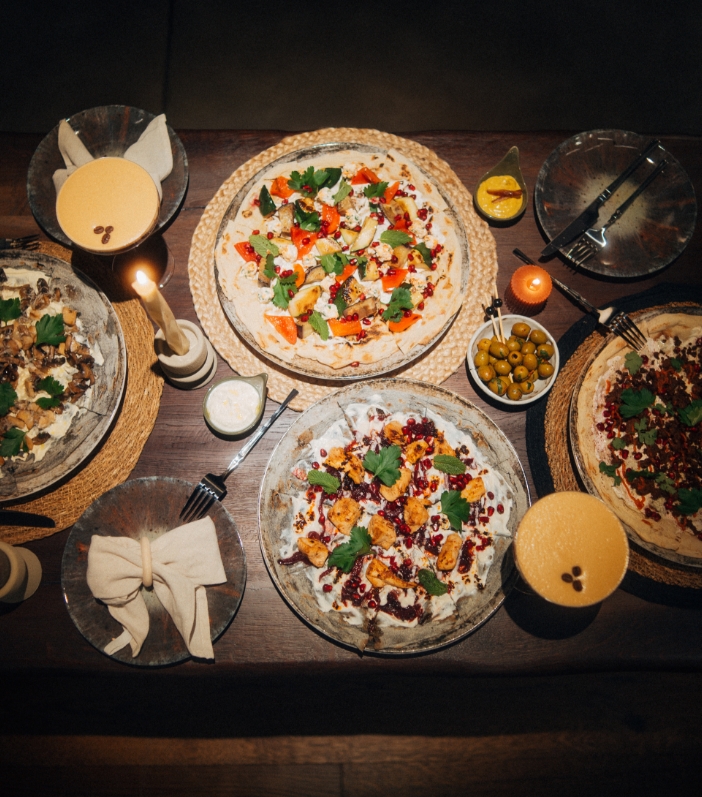 Draufsicht auf einen rustikalen Holztisch, gedeckt mit vier verschiedenen belegten Fladenbroten, zwei gelben Cocktails mit Kaffeebohnen, Oliven und Kerzenlicht in gemütlicher Atmosphäre.