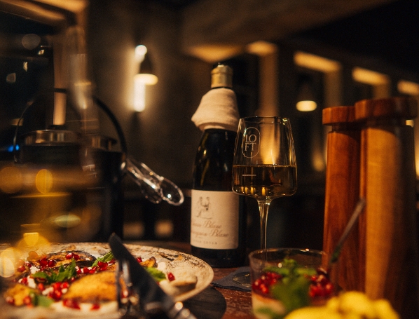 Nahaufnahme eines Weinglases mit Weißwein und einer Weinflasche, die neben einem Teller mit Essensresten (Aubergine/Pizza) und einem Holzgefäß auf einem dunklen Tisch stehen.